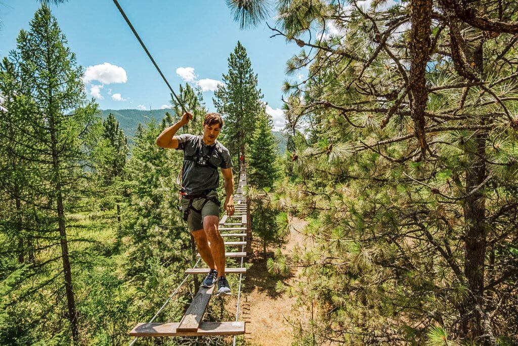 A man walks along a wood plank bridge while holding a rope, surrounded by pine trees.