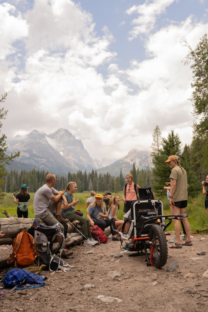 A large group of people are gathered around an all-terrain wheelchair looking at the jagged Sawtooth Mountains.