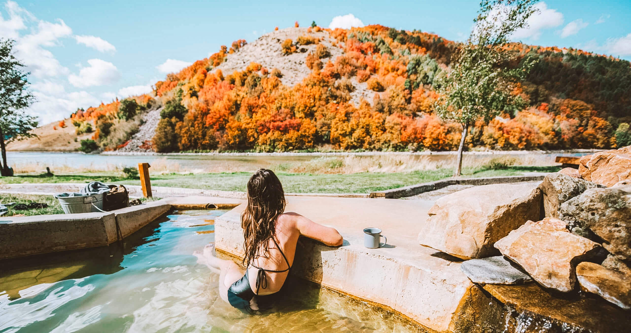 A woman wearing a black one piece bathing suit, sitting in a hot pool, staring at a hill covered in red, orange, yellow and green foliage.