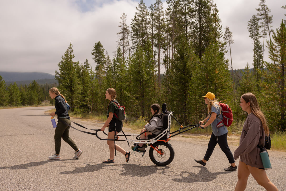 A group of three females assists a woman in an all-terrain wheelchair on an asphalt trail.