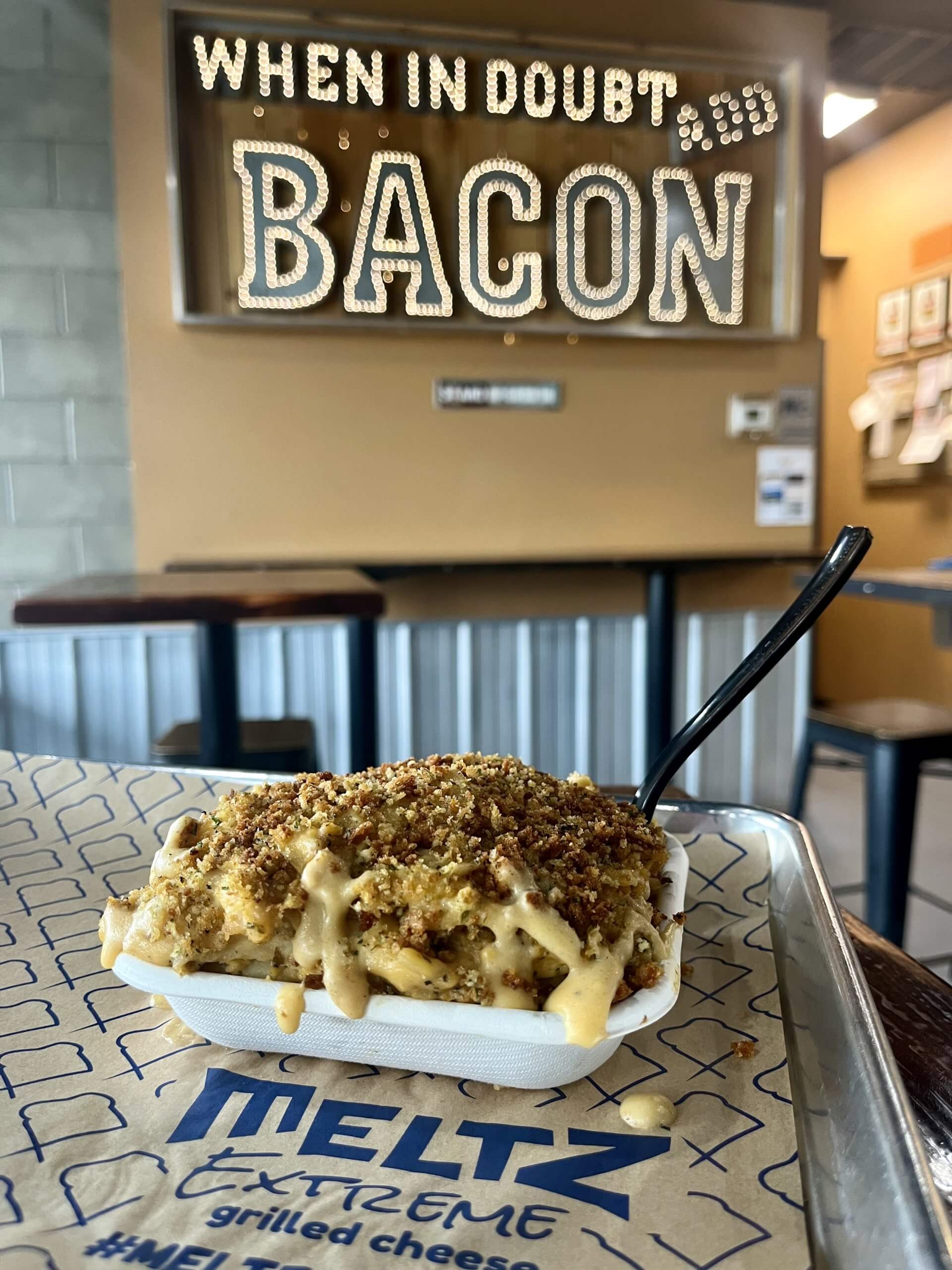 A dish of cheesy scalloped potato pie topped with a crouton crust on a table at Meltz Extreme, and a lit up sign that reads, "When in Doubt, Add Bacon" in the background.