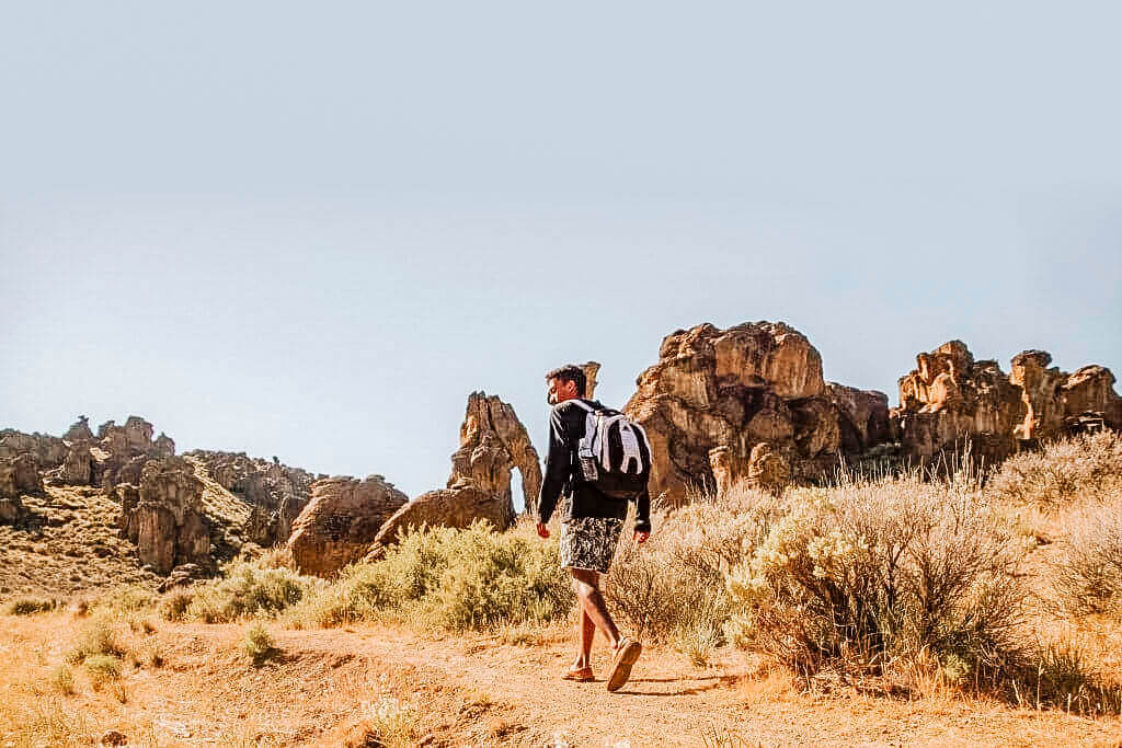 A man in summer hiking gear walks toward earth-colored rock formations.