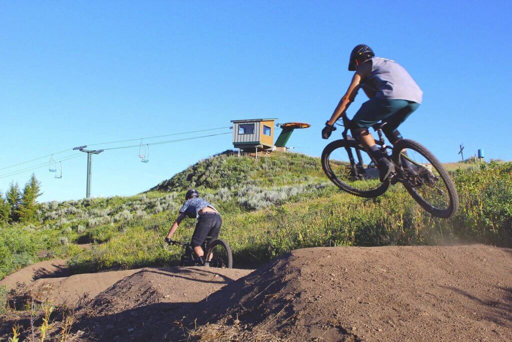 Two people riding mountain bikes over a series of jumps beside a field.