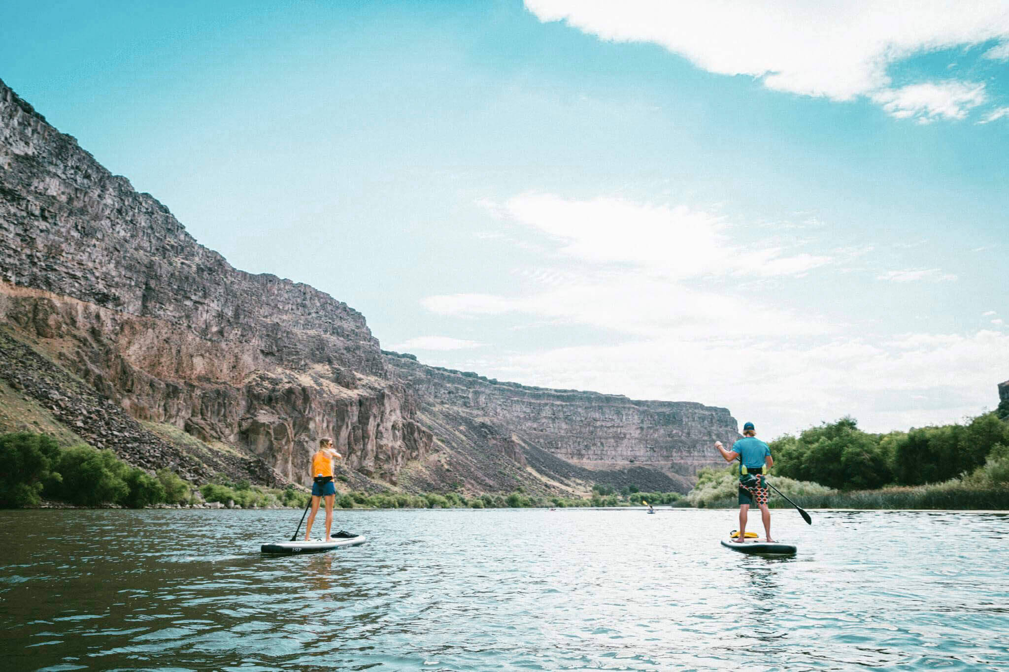 Two people stand-up paddleboarding on the Snake River past a rugged landscape.