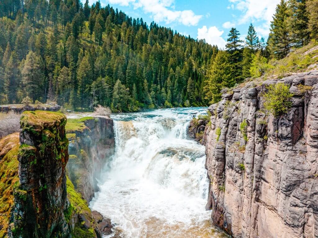 Lower Mesa Falls flowing between two large walls of rock and a forest of trees in the distance.