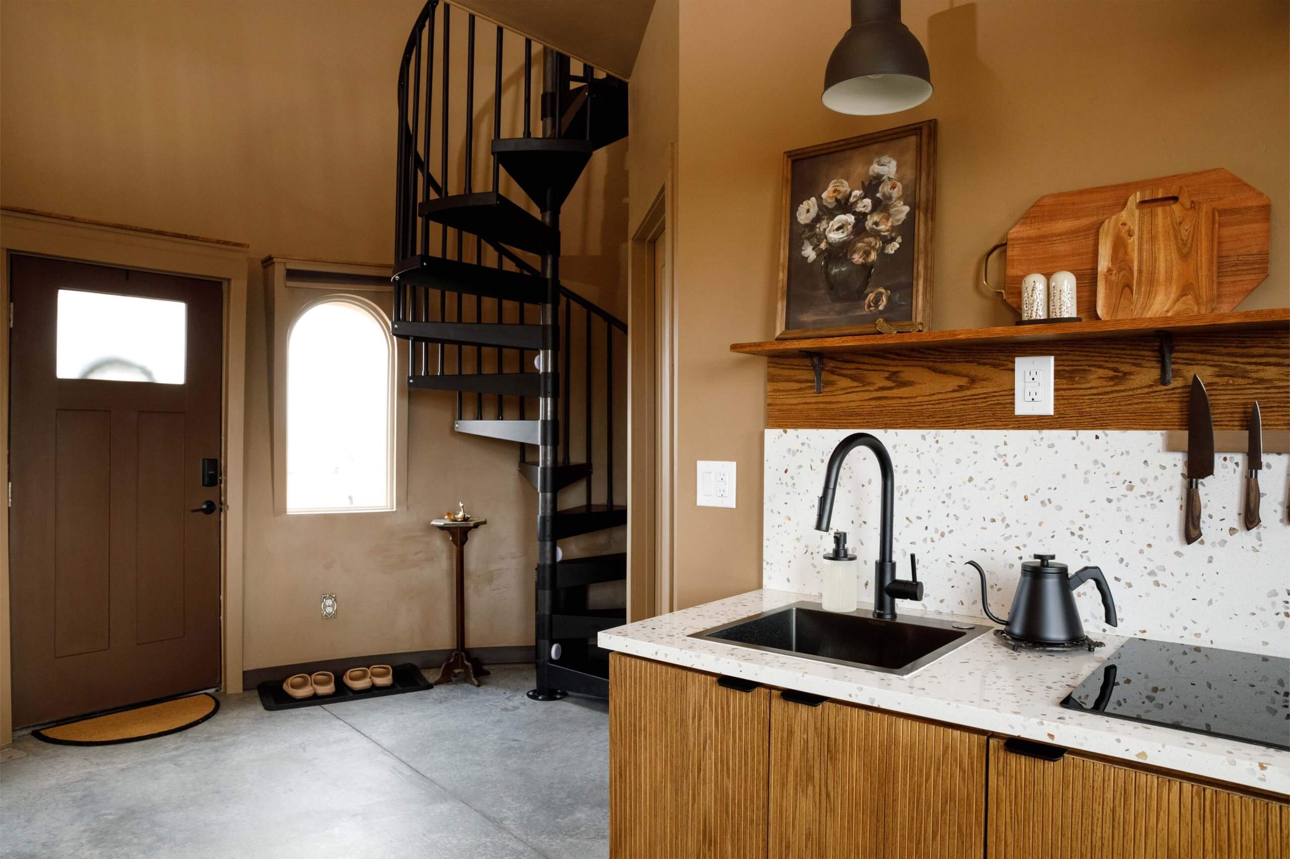 A view of the kitchen, where a framed photo of flowers hangs above the faucet as a spiral staircase beckons in the distance before a glowing window.