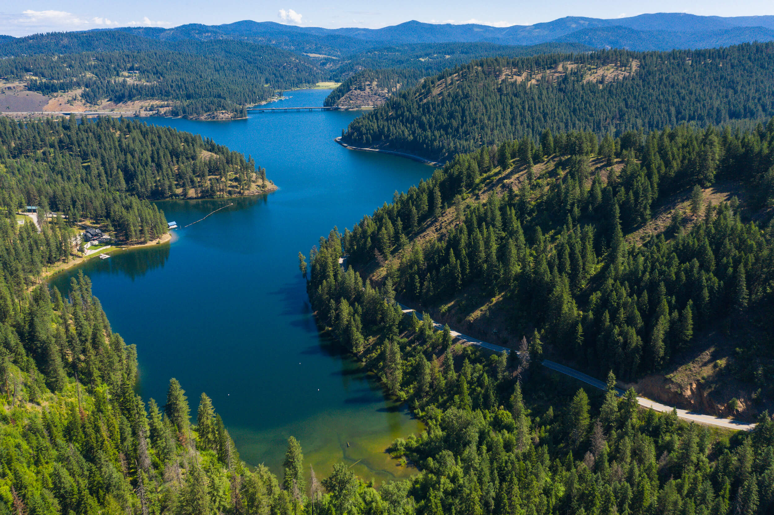 An aerial view of Lake Coeur d'Alene lined by forests of trees and the Lake Coeur d'Alene Scenic Byway winding alongside it.