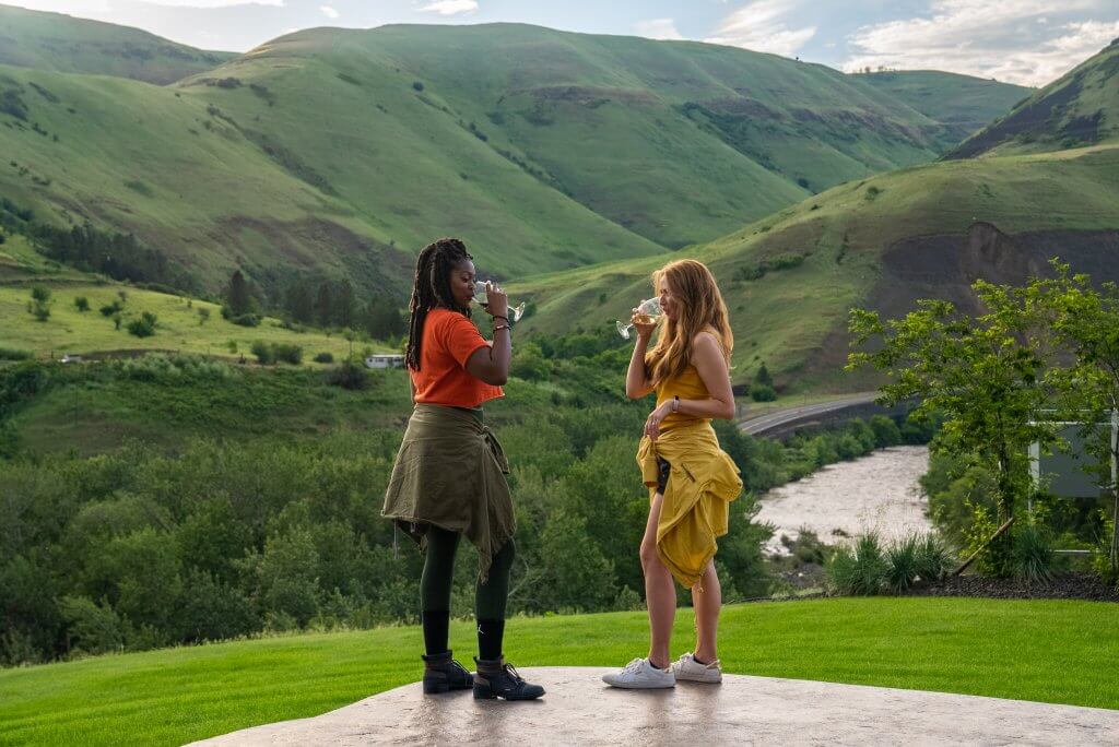 Two women sipping wine on a patio and landscape of green hills and mountains in the background at Rivaura Estate Vineyard & Winery.