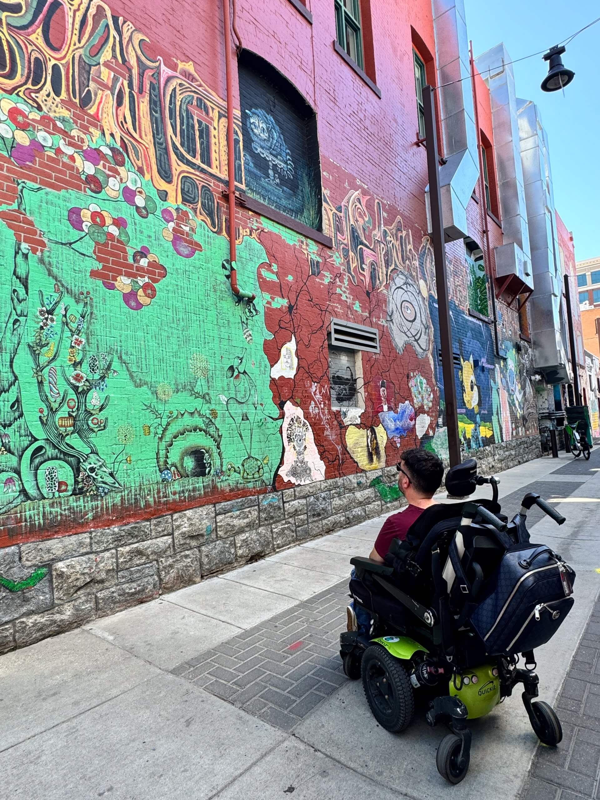 A man in a wheelchair sits looking at a brick wall covered in vibrant painted murals.