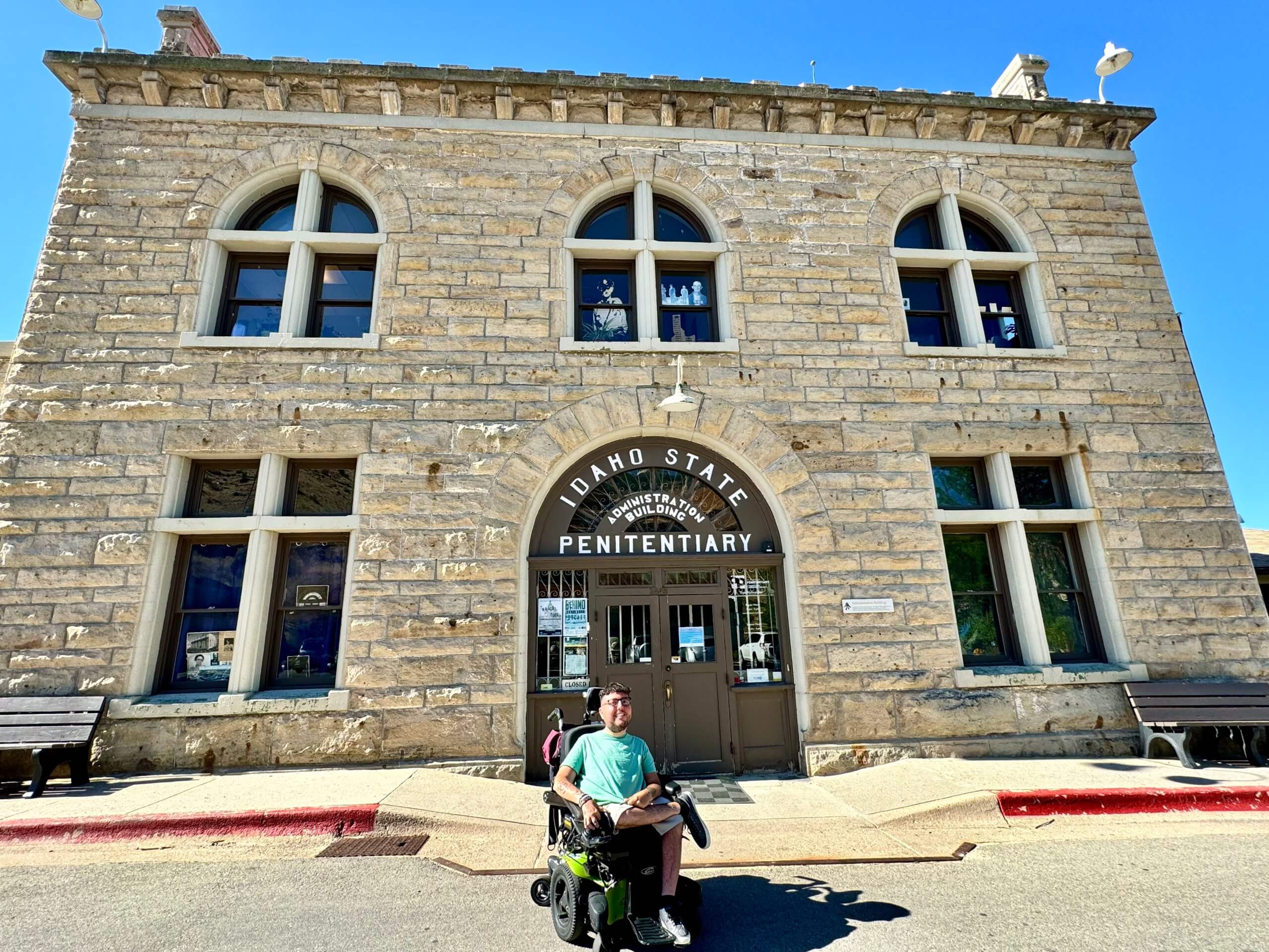 A man in a wheelchair sits in front of the light brown brick front of the Old Idaho Penitentiary. 