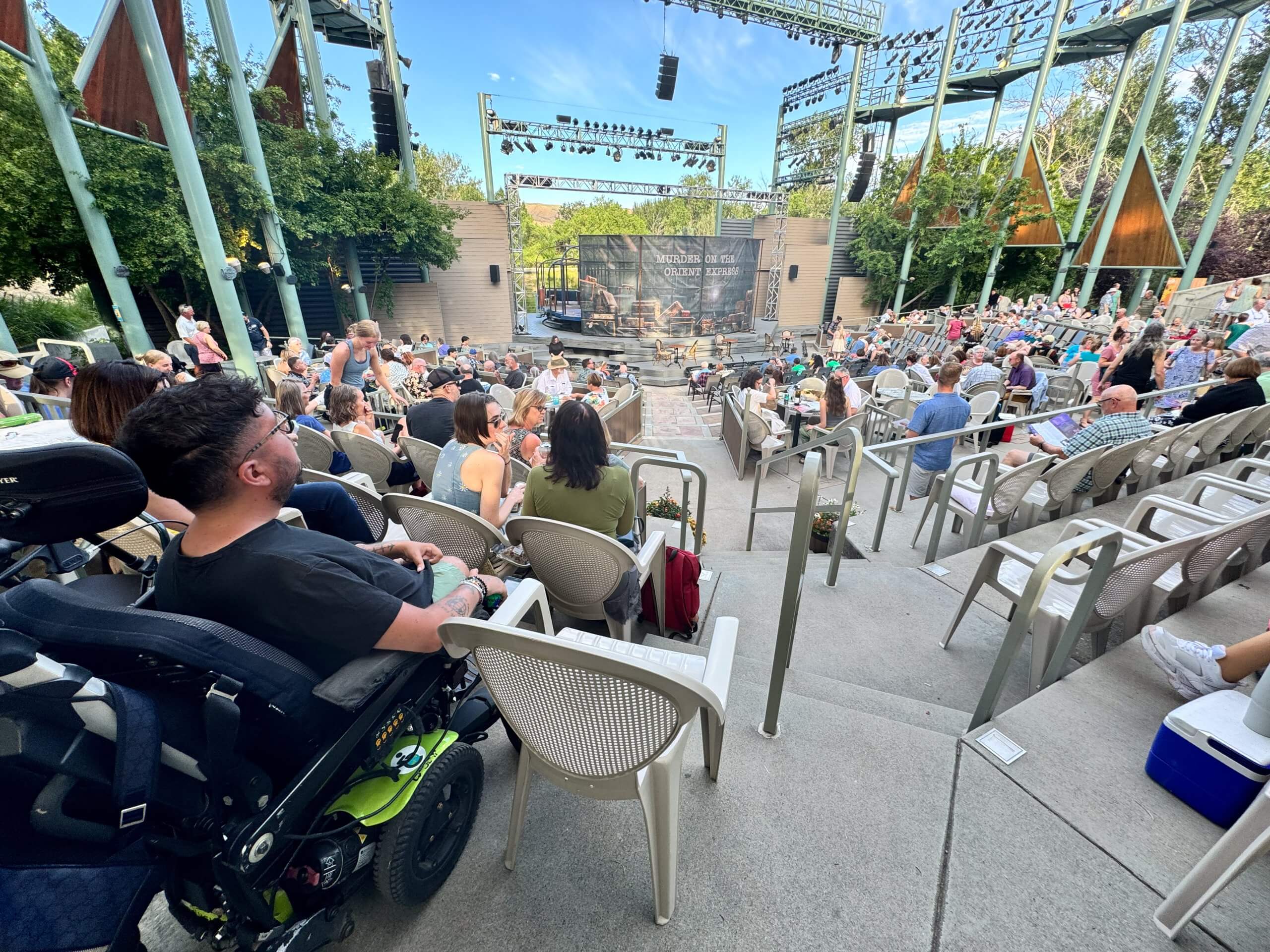 A man in a wheelchair sits in a raised seating area looking at an outdoor amphitheater stage.