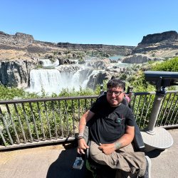 A man in a wheelchair smiles at the camera with the giant Shoshone Falls in the background.