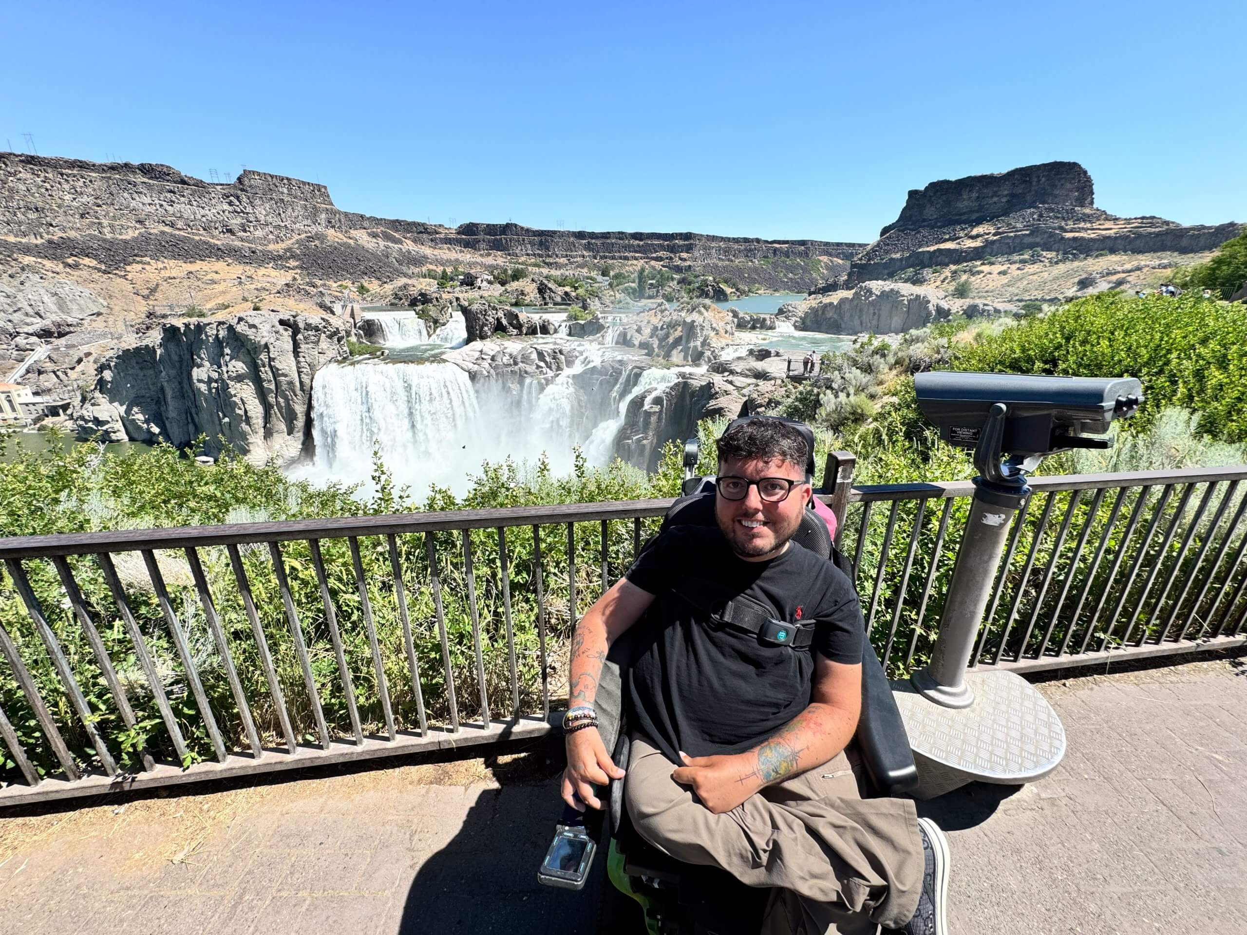 A man in a wheelchair smiles at the camera with the giant Shoshone Falls in the background.