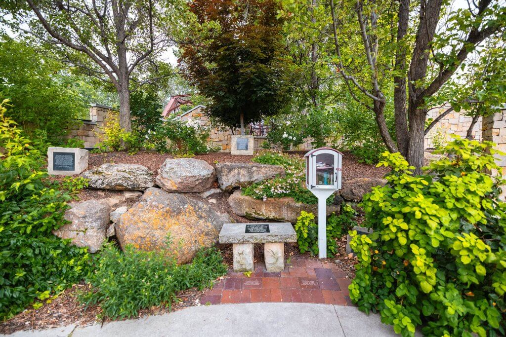 A stone bench with a plaque and a wooden book box surrounded by trees and bushes at the Idaho Anne Frank Human Rights Memorial.