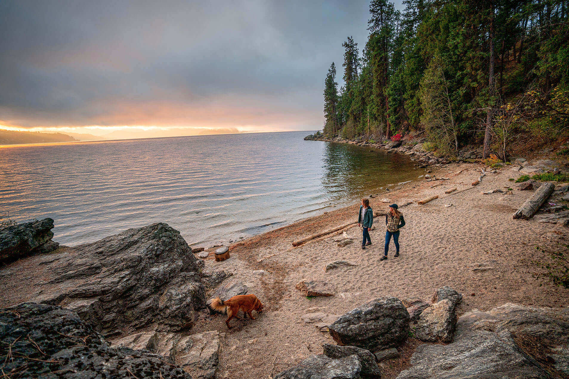 Two people walking along the beach at Tubbs Hill at sunset and a forest of trees in the background.