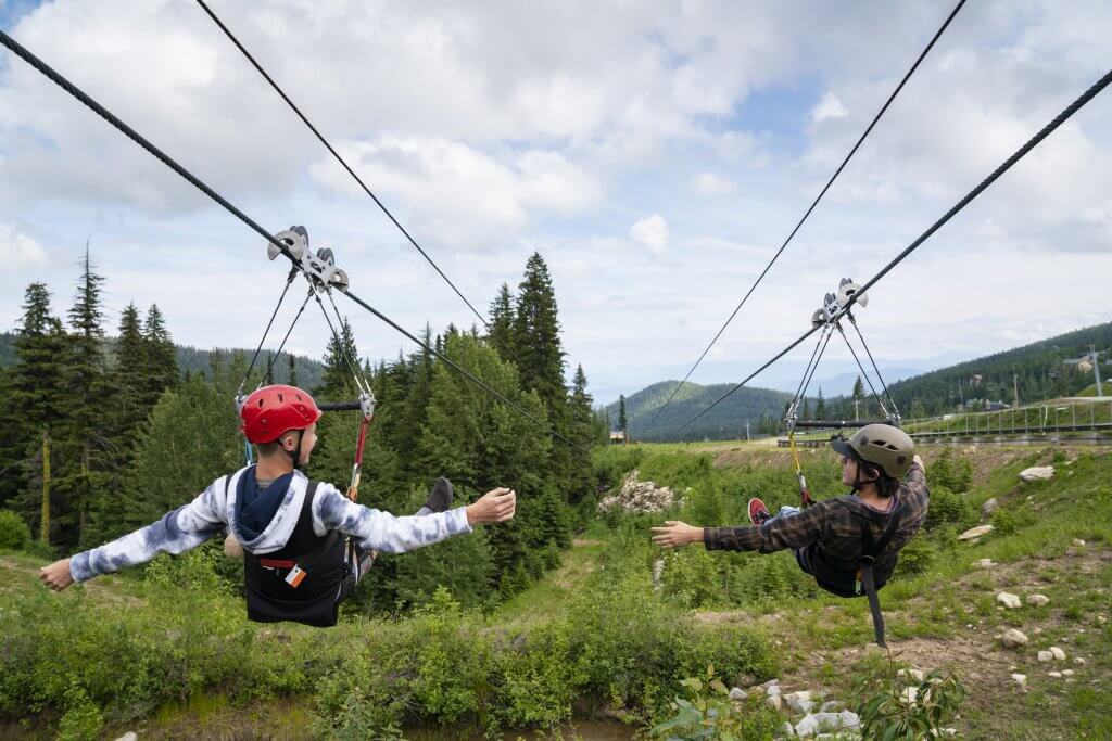 Two children ziplining side by side on tandem ziplines past a forested area at Schweitzer.