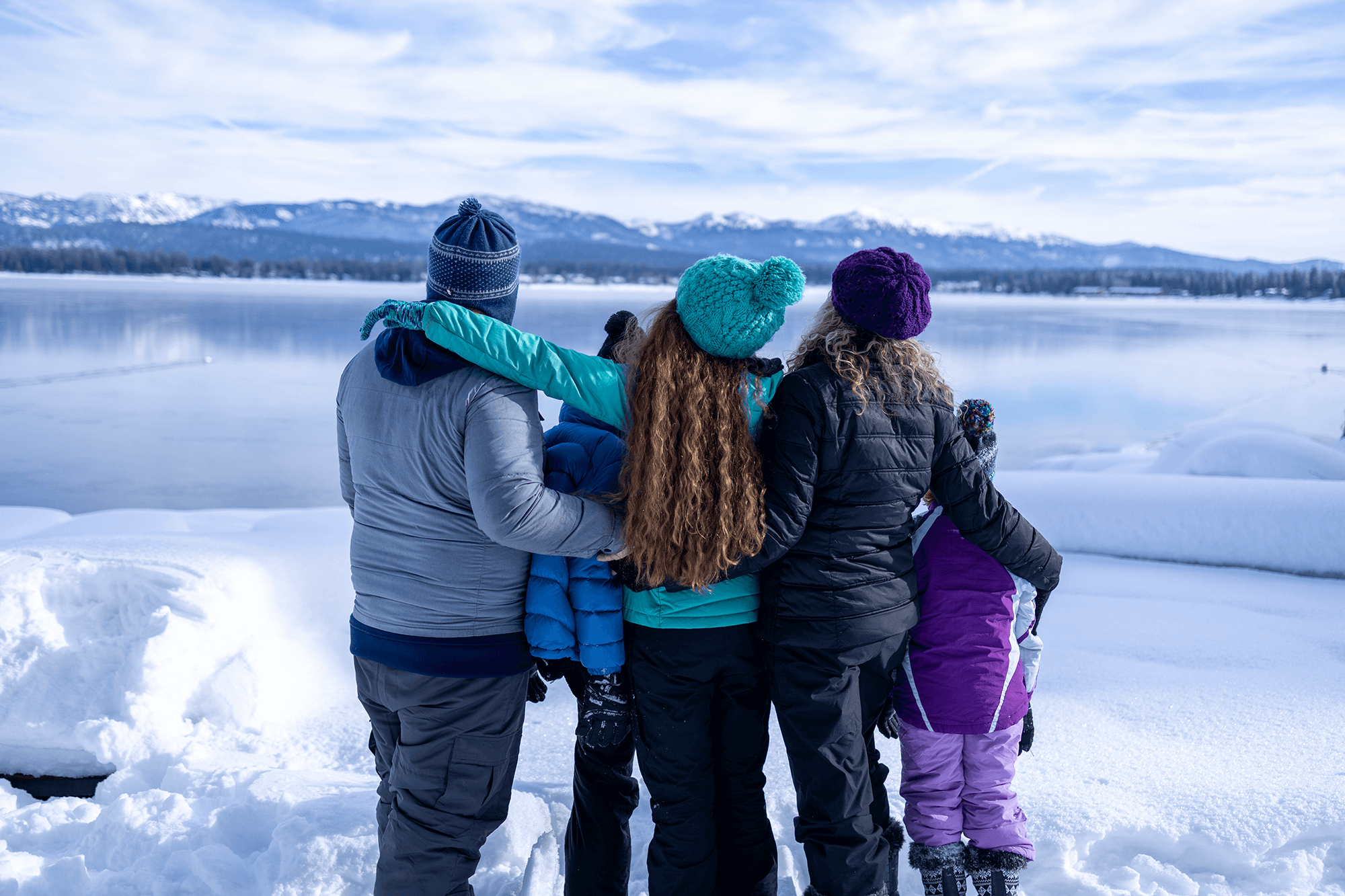 Family of four standing together with their arms around each other, looking out over the snowy landscape of Payette Lake, with mountains in the distance under a bright blue sky.