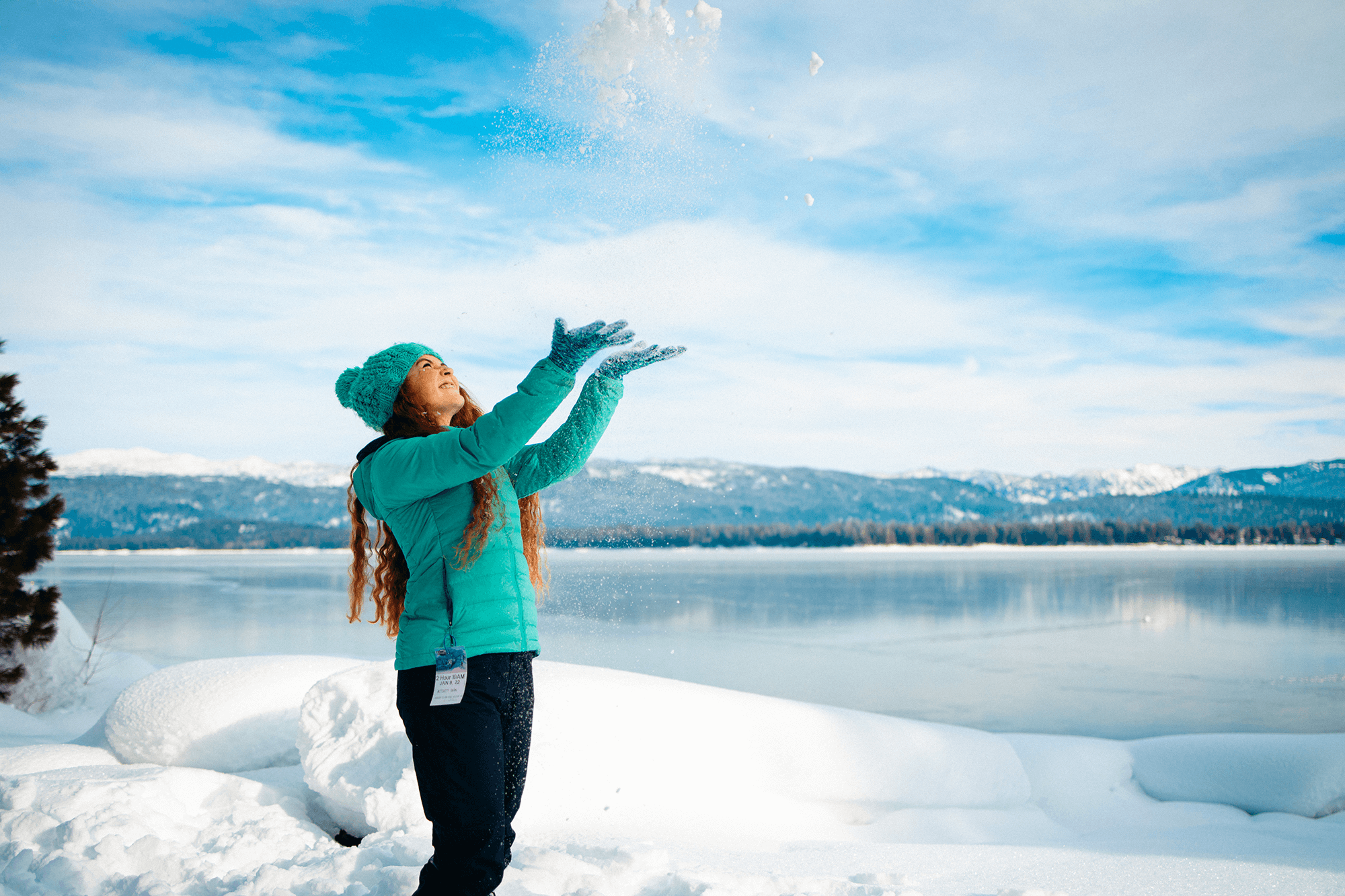 Person in a teal jacket and beanie joyfully tossing snow into the air at the snowy shoreline of Payette Lake, with mountains and a blue sky in the background.