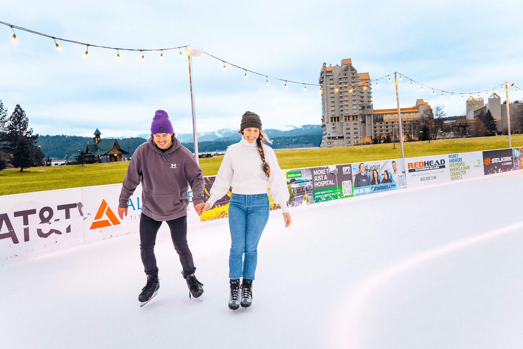 Two people ice skating outdoors on a rink surrounded by festive string lights, with buildings, a lodge, and snowy hills in the background.