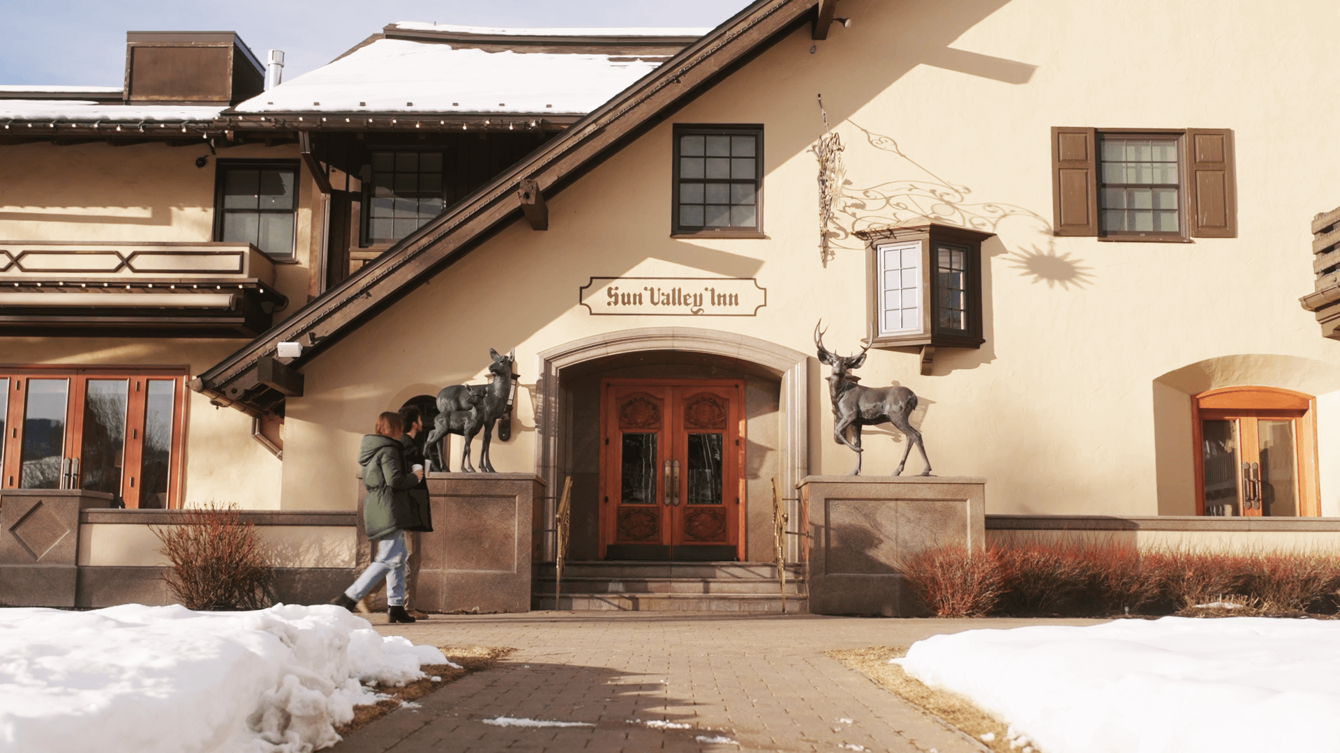 Two people walking past the Sun Valley Inn and a snowlined walkway out front.