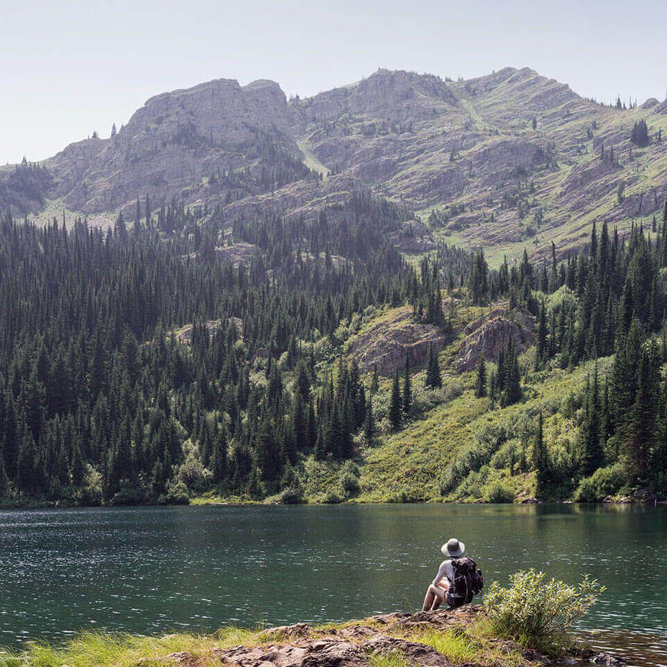 A backpacker rests before a sparkling lake on Stevens Lakes Trial, looking up at a mountain lined with trees.