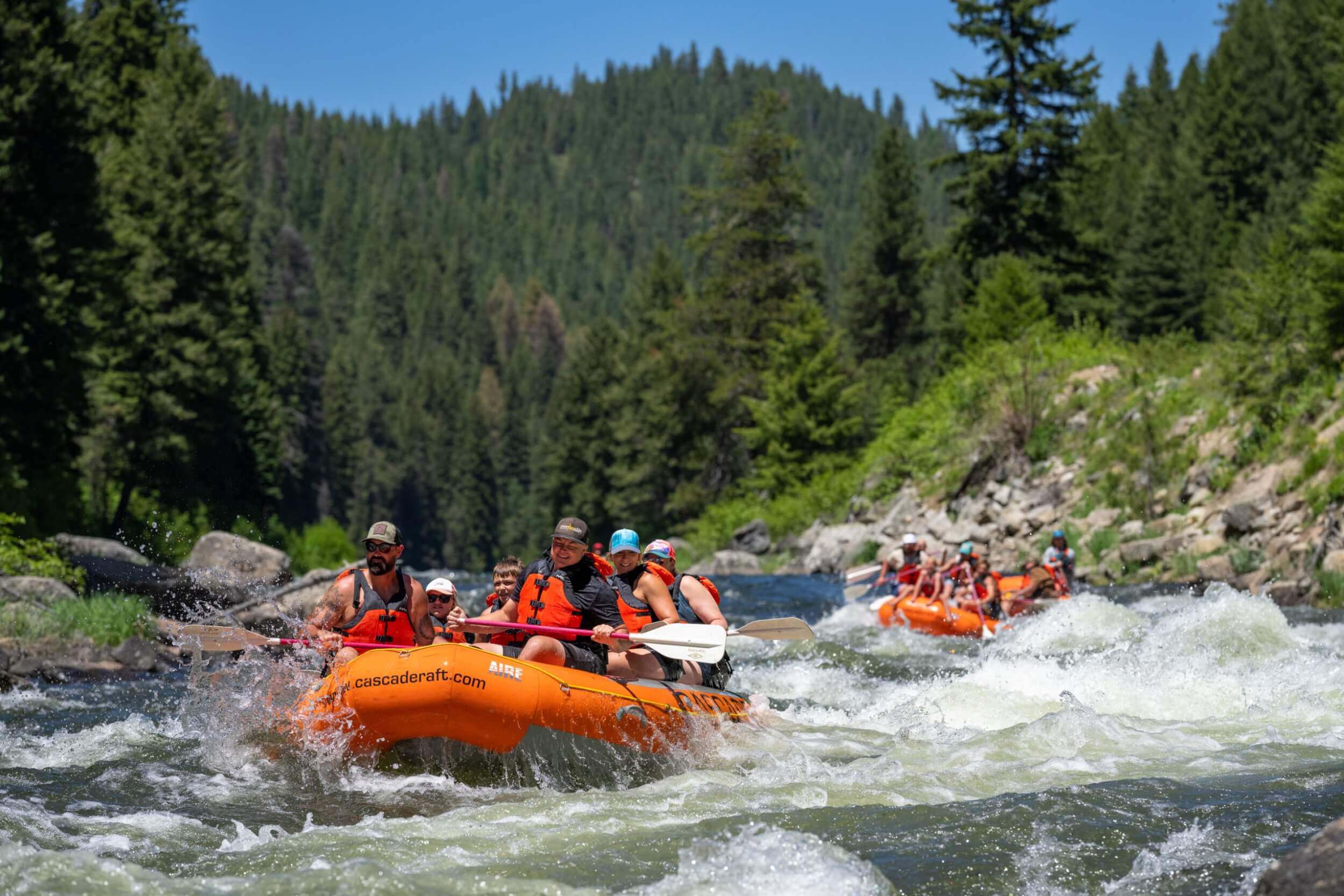 A group of smiling rafters paddle through gentle rapids in a bright orange Cascade Raft & Kayak boat.