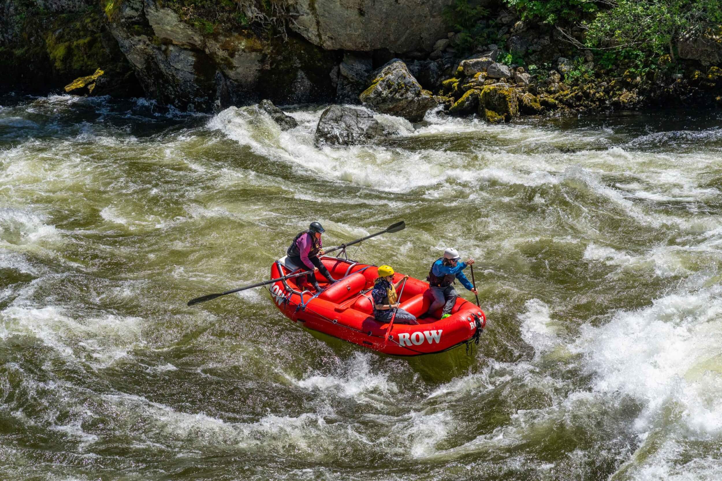 Three people paddle a red ROW Adventures raft through whitewater rapids