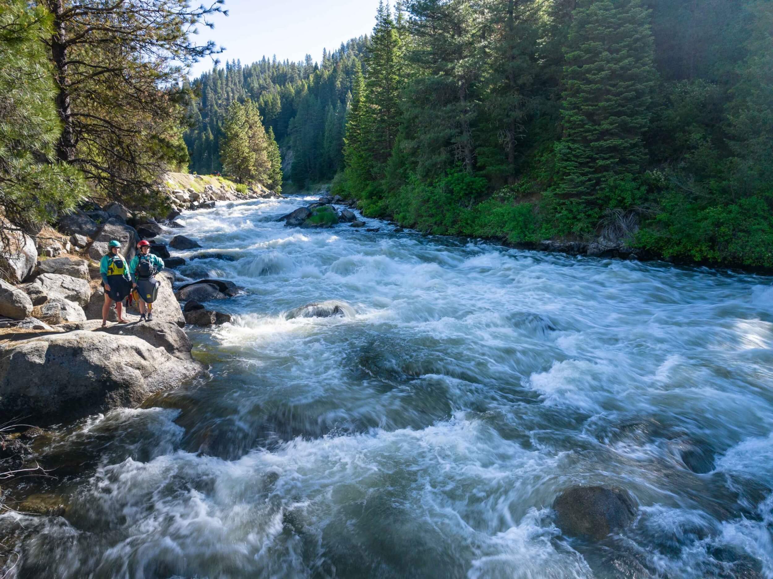 Sage Donnelly, Professional Kayker & Raft Guide and Darby McAdams, Professional Kayaker & Filmmaker stand on rocks beside a rushing section of the Payette River, scouting the rapids.