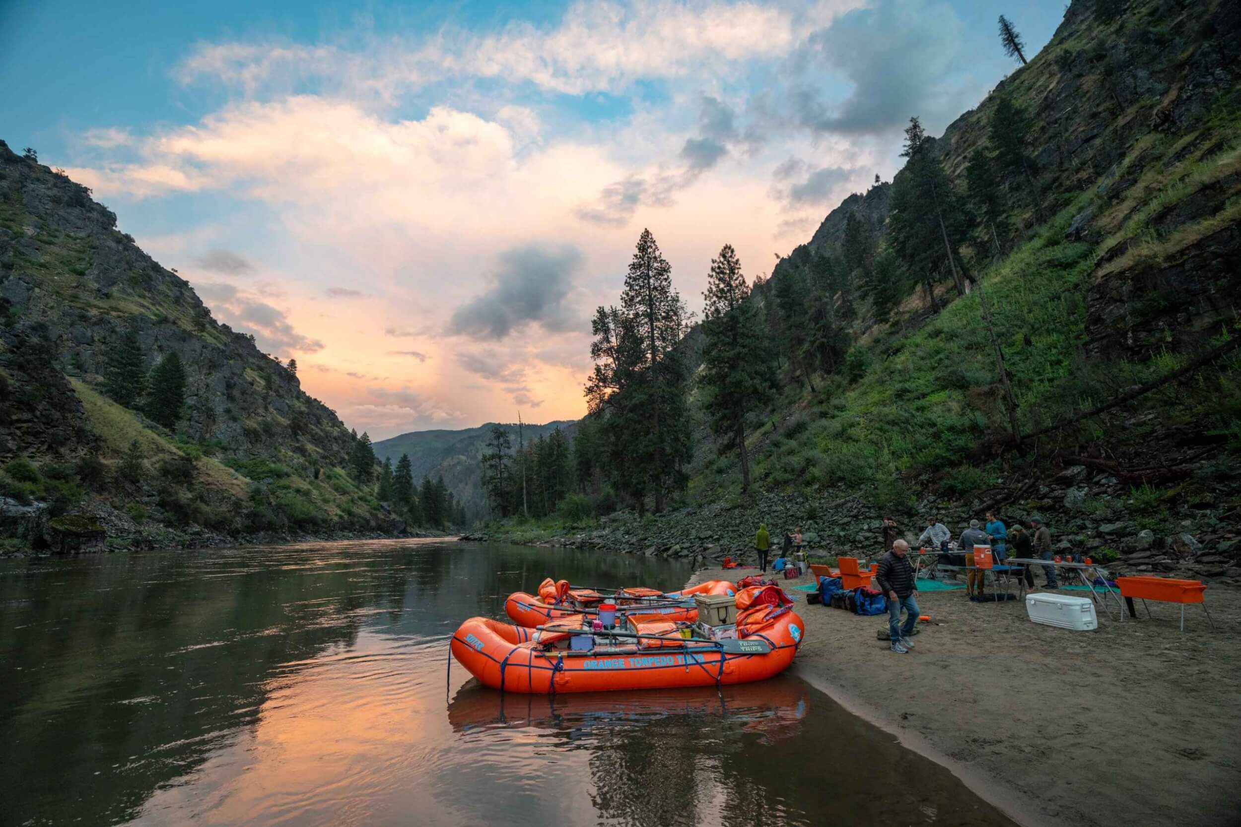 Rafts docked on a sandy beach at sunset on the Main Salmon River with people preparing camp.