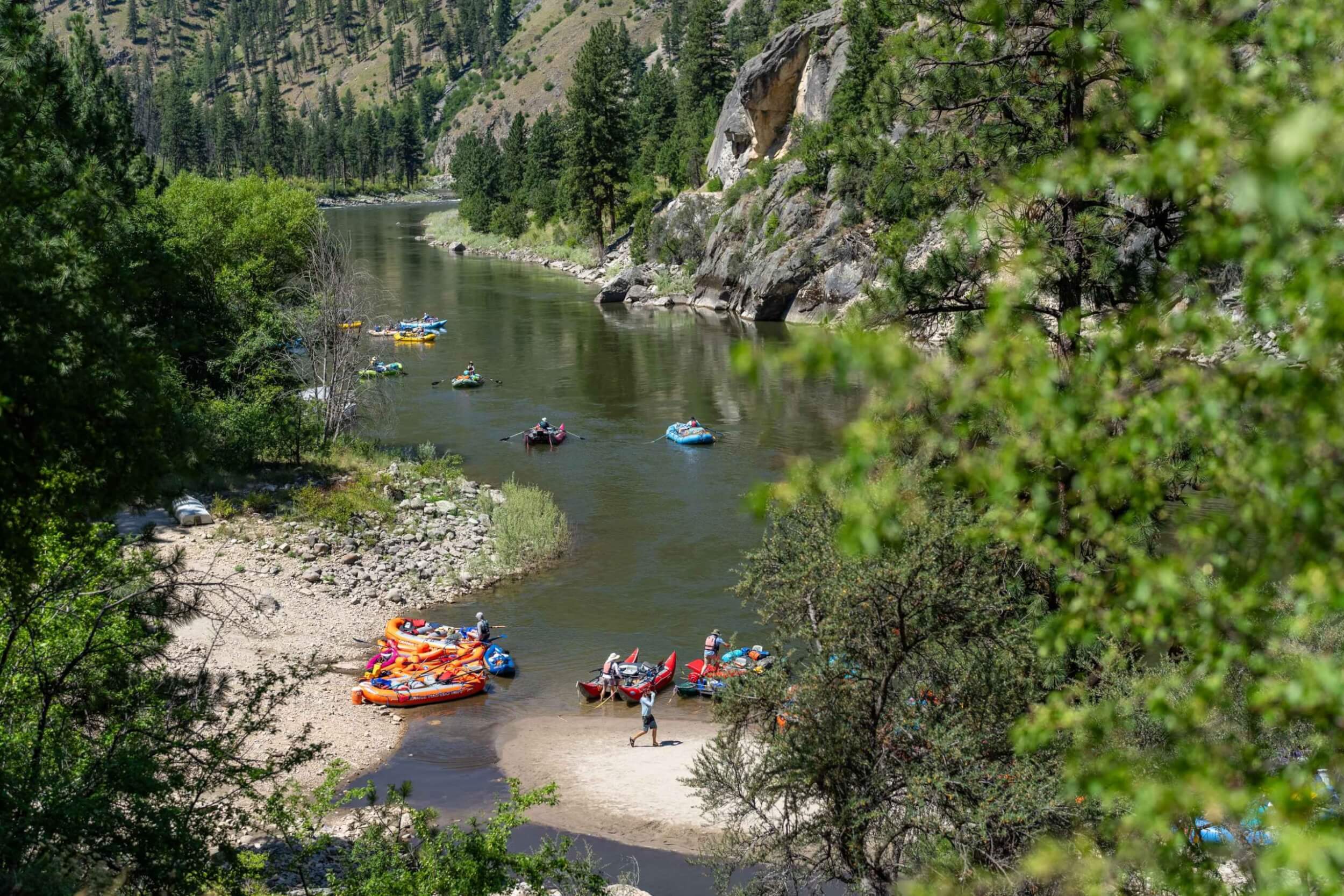 Rafts and inflatable kayaks gathered at a sandy beach while more float down the river.