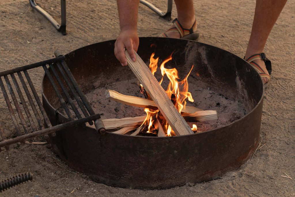 TRAVEL WITH CARE: CAMPFIRE SAFETY A person adds a piece of wood to a campfire enclosed in a ring.