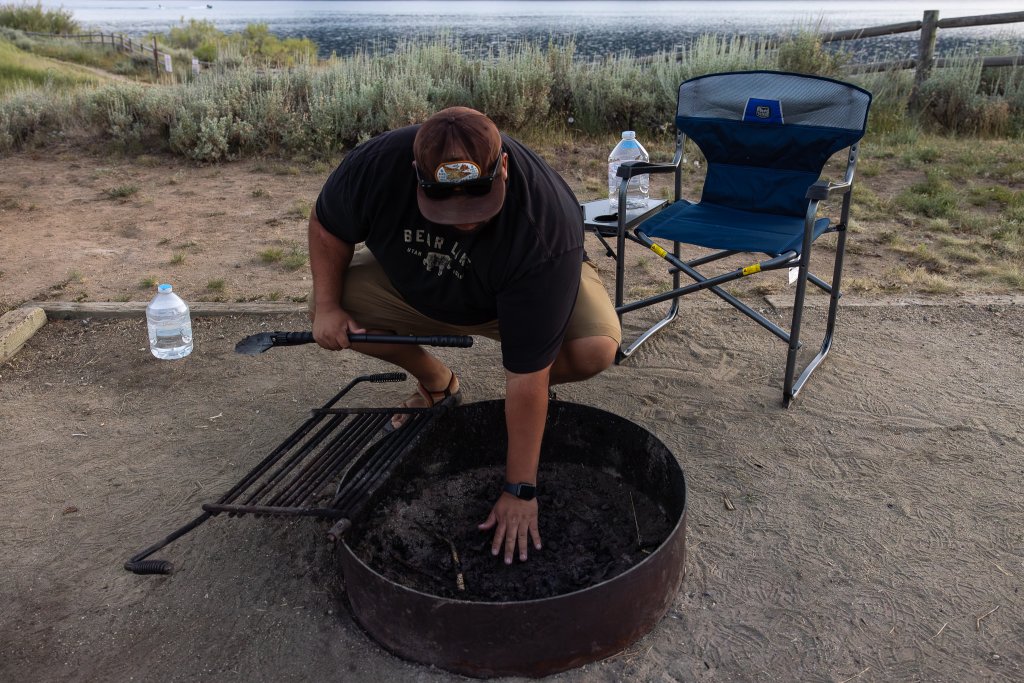 TRAVEL WITH CARE: CAMPFIRE SAFETY A camper ensures their campfire is completely out by holding their hand above the coals, making sure they are cool.