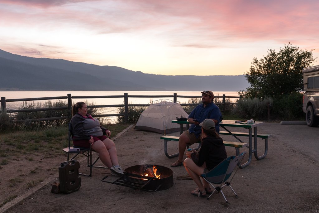 TRAVEL WITH CARE: CAMPING WITH CARE Three campers sit around a campfire at sunset next to a lake.