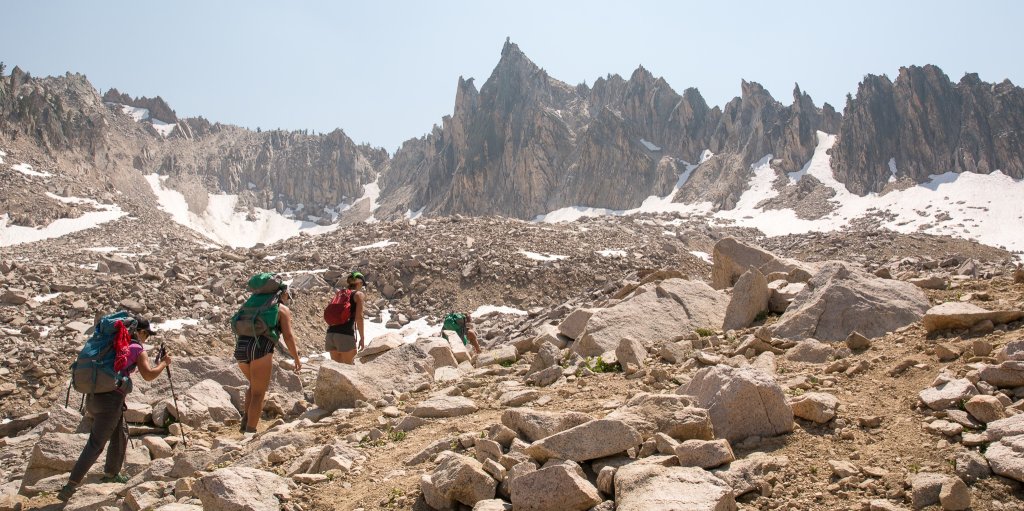 Hikers follow a rocky trail to a snowy summit.