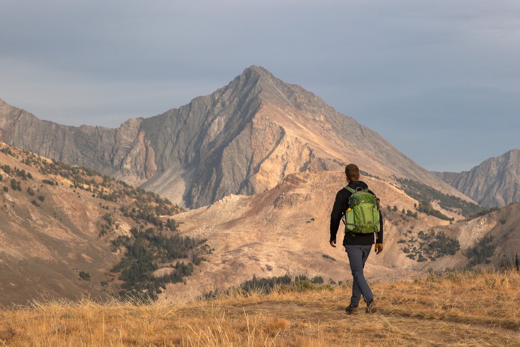 TRAVEL WITH CARE: HIKING RESPONSIBLY A hiker carrying a green backpack walks along a trail with a mountain in the distance.