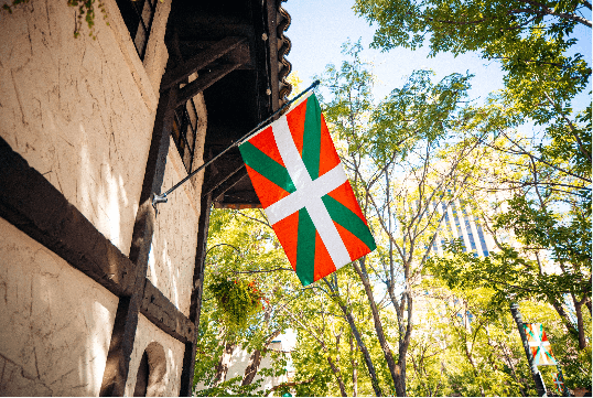 A Basque flag flying hanging on the outside wall of the Basque Center, and in the background, several trees and a tall building.