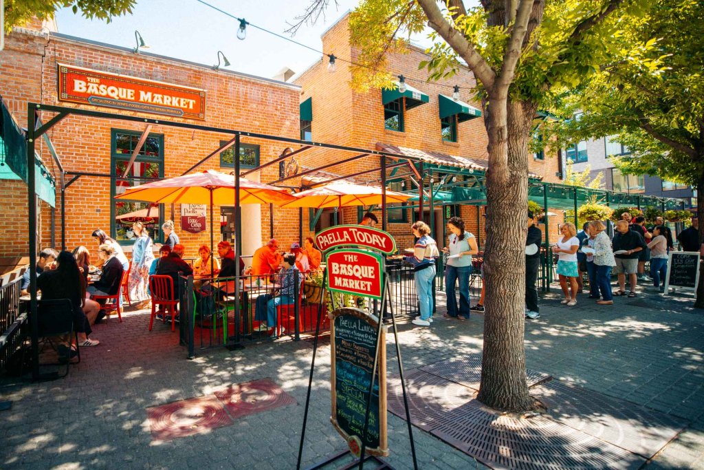 An outdoor view of The Basque Market brick building and a group of people sitting beneath umbrellas on the patio out front.