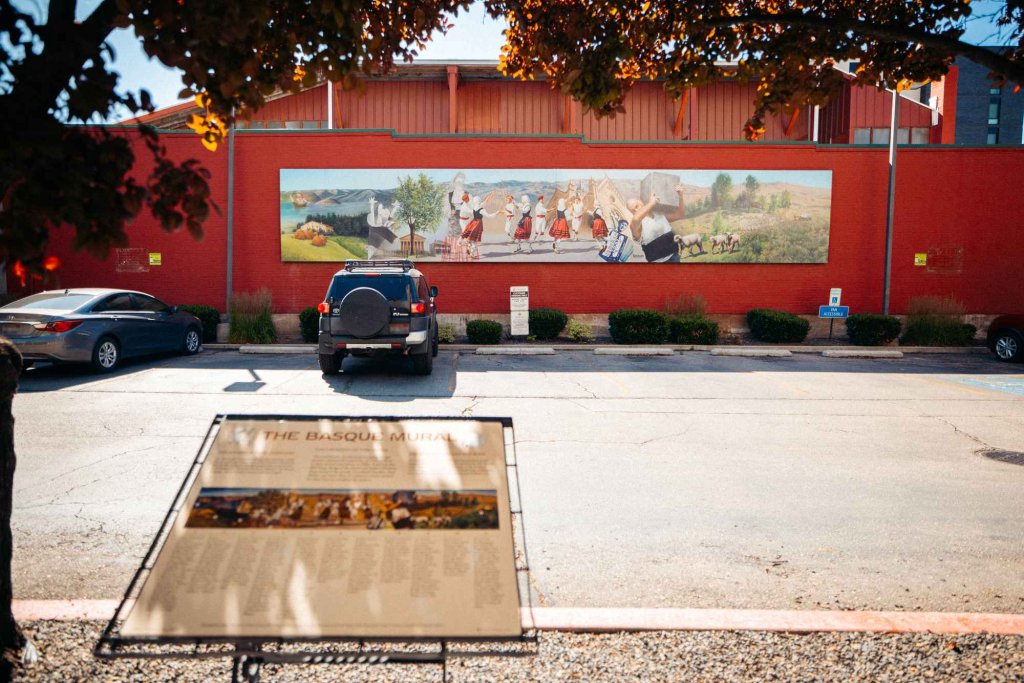 In the foreground, an interpretive sign for the Basque Mural. And in the background, two cars in a parking lot in front of the Basque Mural depicting Basque dancers, grazing sheep, scenery and a man carrying a hay bale.