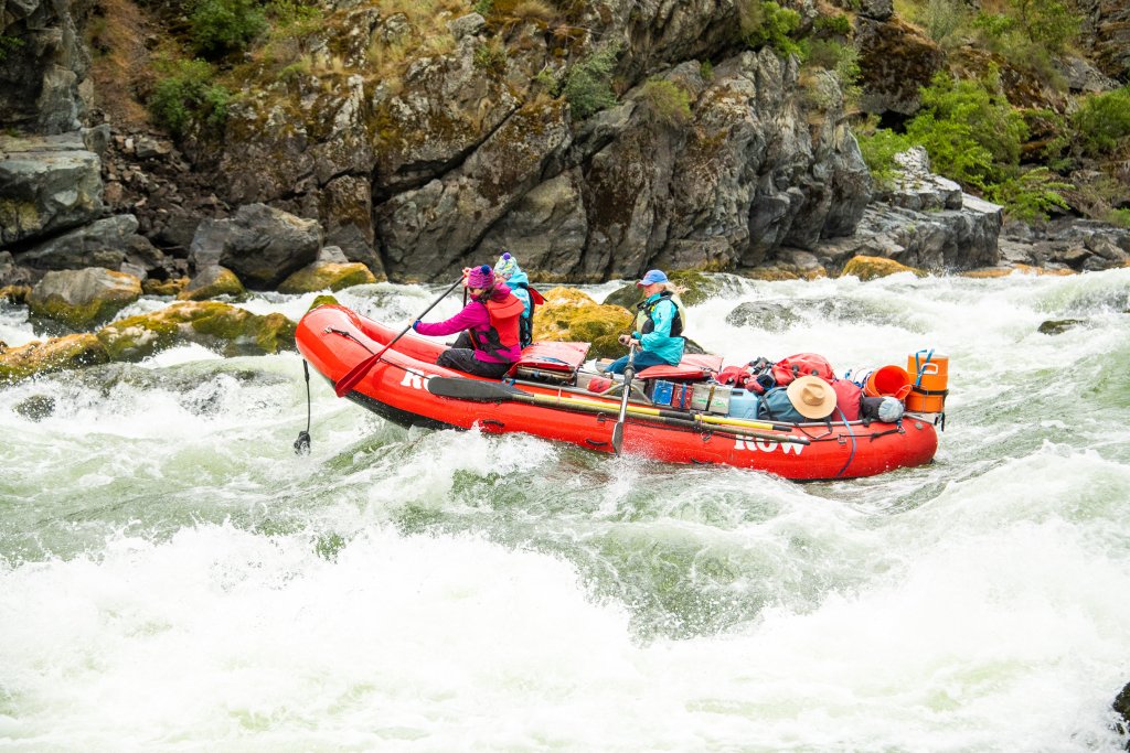 Meet the Queen of the Snake River Three people wearing life jackets sitting on an orange raft paddle through a rapid on the Snake River.