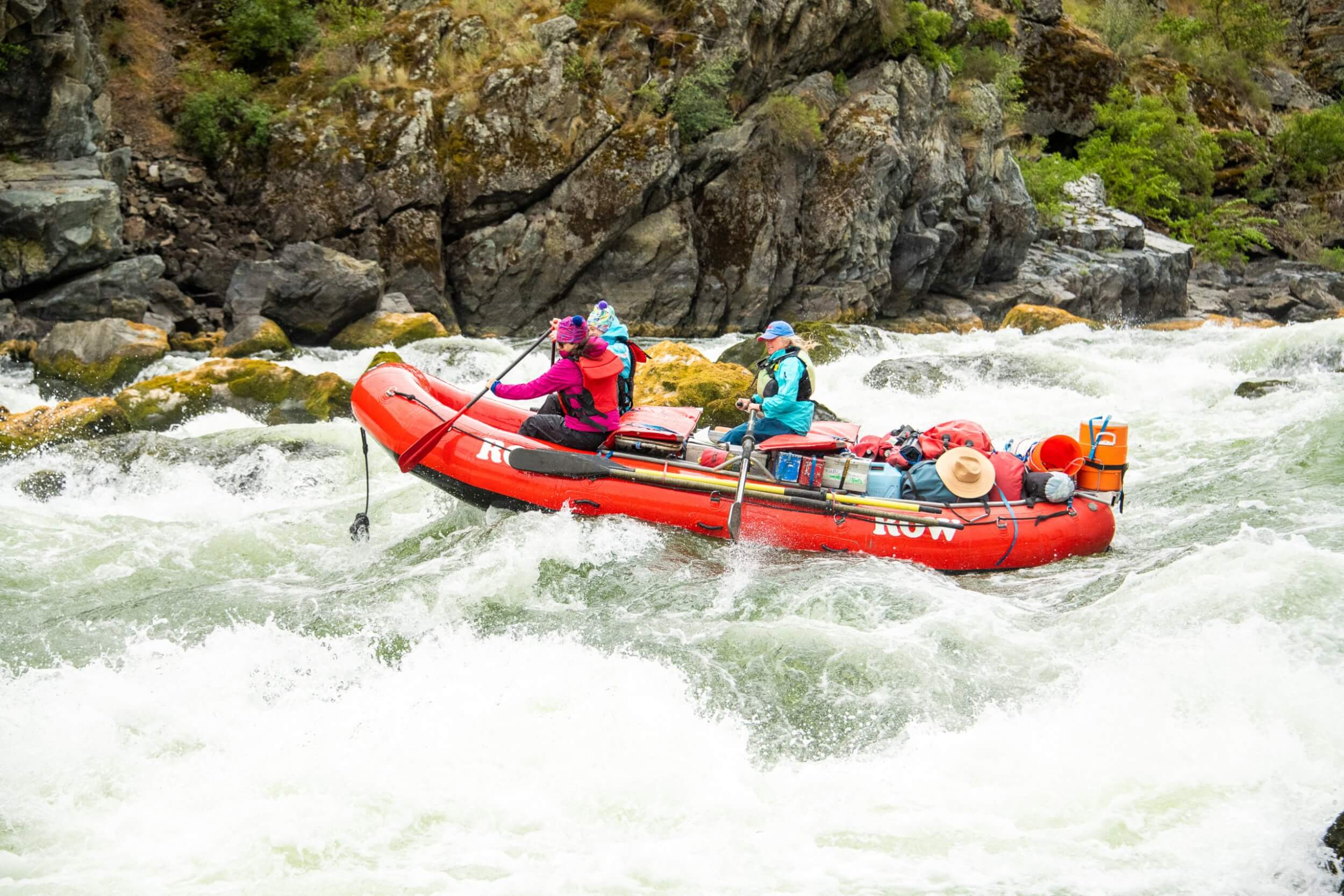 Meet the Queen of the Snake River Three people wearing life jackets sitting on an orange raft paddle through a rapid on the Snake River.