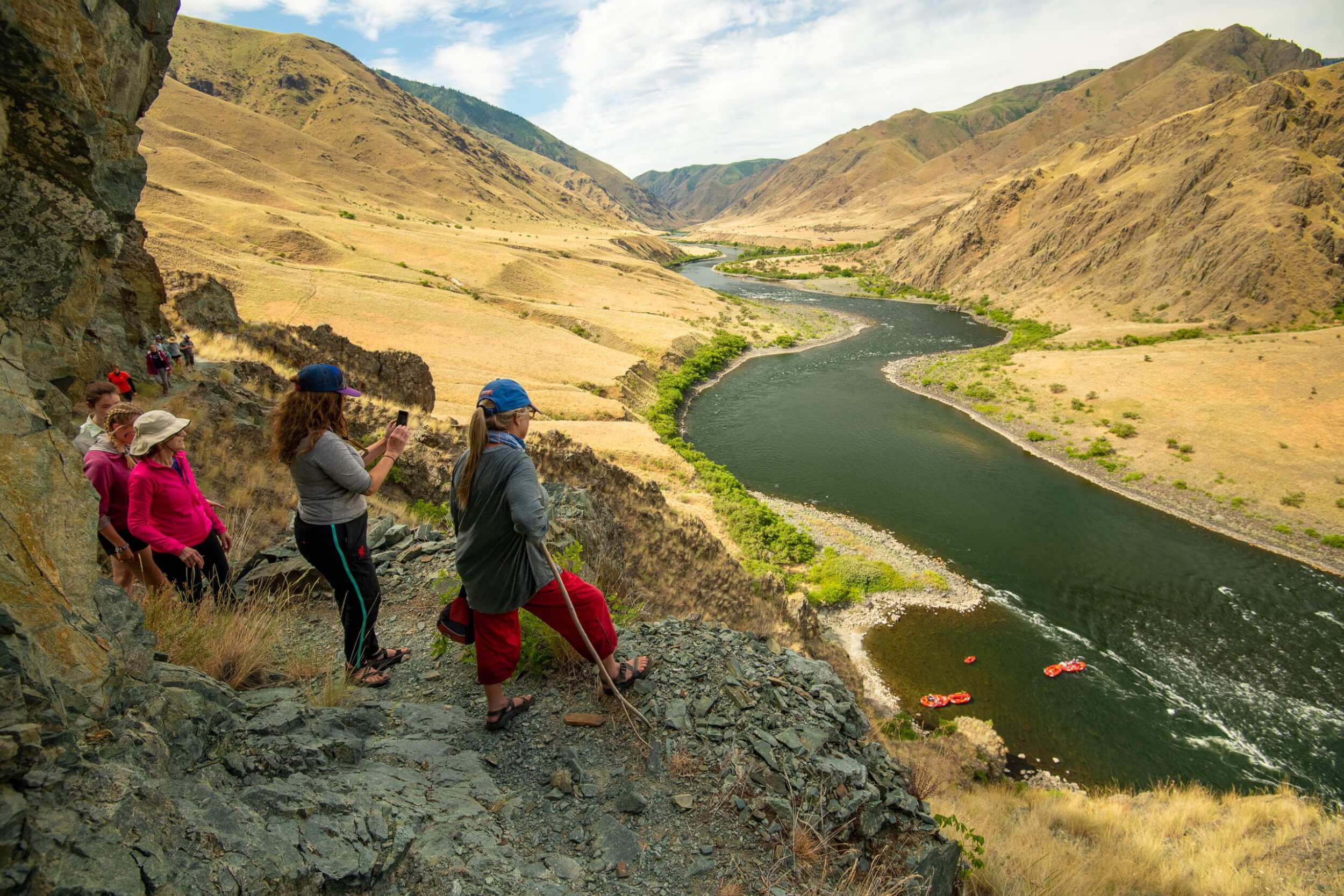 Meet the Queen of the Snake River A group of people stand on a rock ledge looking down to the Snake River where four orange whitewater rafts are anchored.