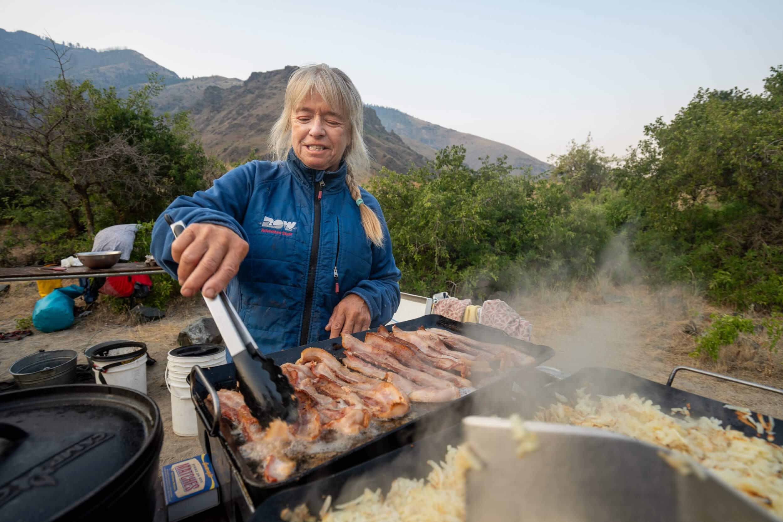 Meet the Queen of the Snake River A woman in a blue coat is using tongs to flip bacon on an open-air flat top grill.