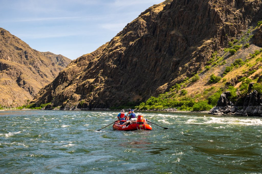 Meet the Queen of the Snake River Three people sit on an orange raft looking down stream as the oar are in some rapid ripples on the Snake River.