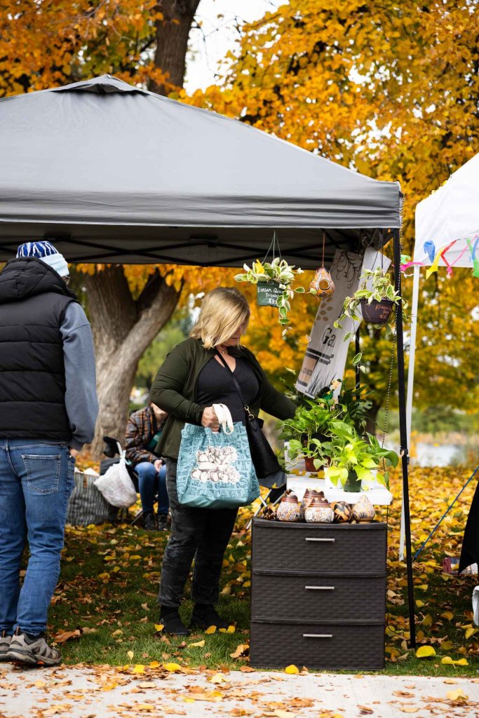 Woman shopping for plants at a fall farmers market booth under golden trees in Idaho Falls.