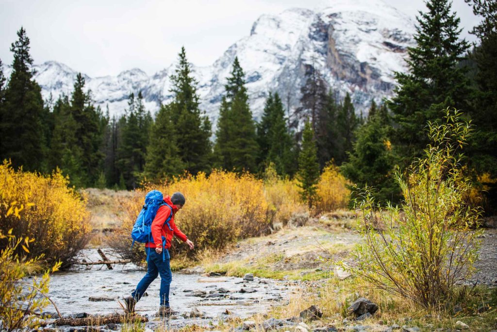 Hiker in red jacket crosses a rocky stream surrounded by fall brush and snow-capped peaks.