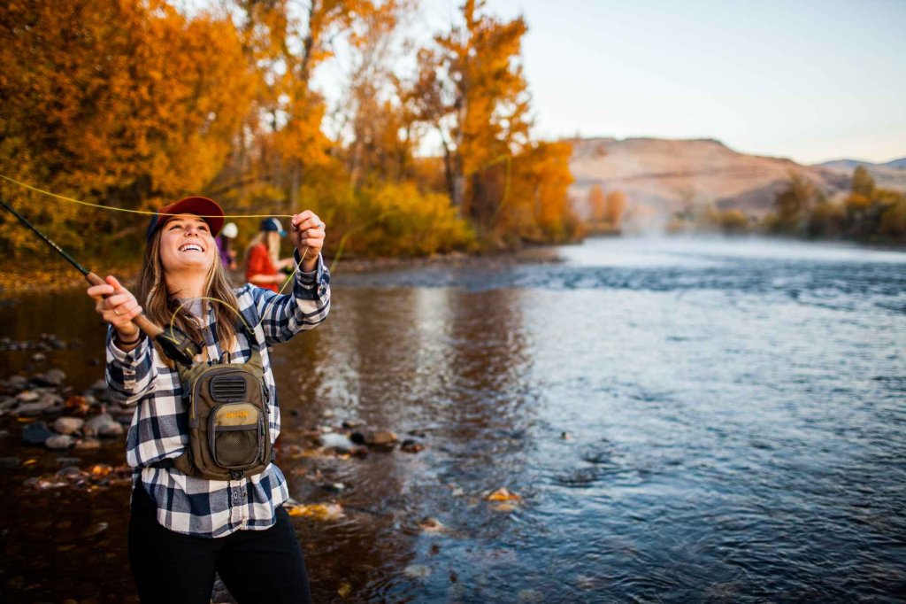 A woman fly-fishing in the Boise River, and a forest of trees with fall foliage in the background.
