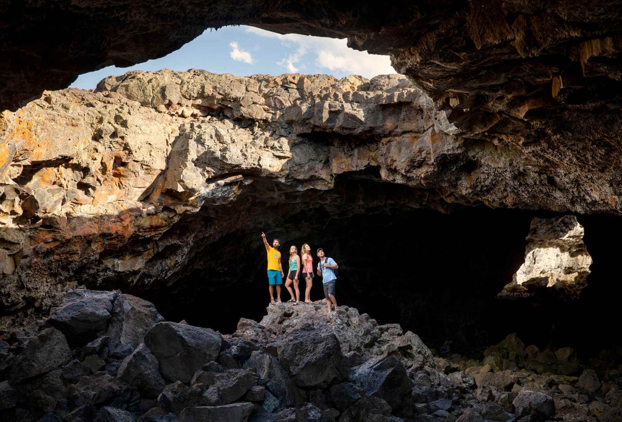A family marvels inside a rugged Craters of the Moon cave, with the cloudy blue sky peeking above.