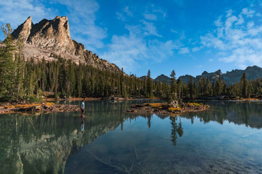 A person stands above Alice Lake as the sky reflects in the water, with vast mountains and forests stretching into the distance.