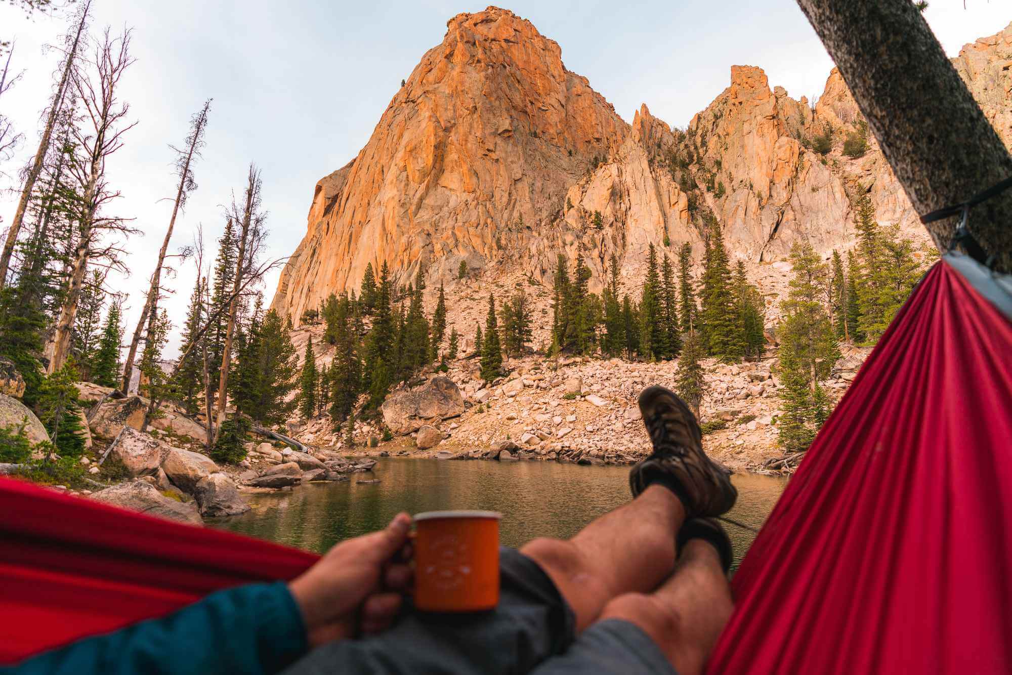 A person lounges in a red hammock with hiking boots up, holding a coffee mug while looking at the Sawtooth peaks over Saddleback Lake.
