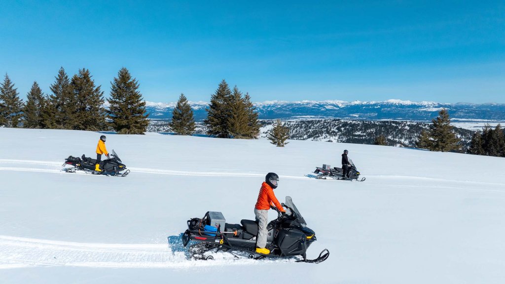 Group snowmobiling across a wide open snow-covered ridge with mountain views